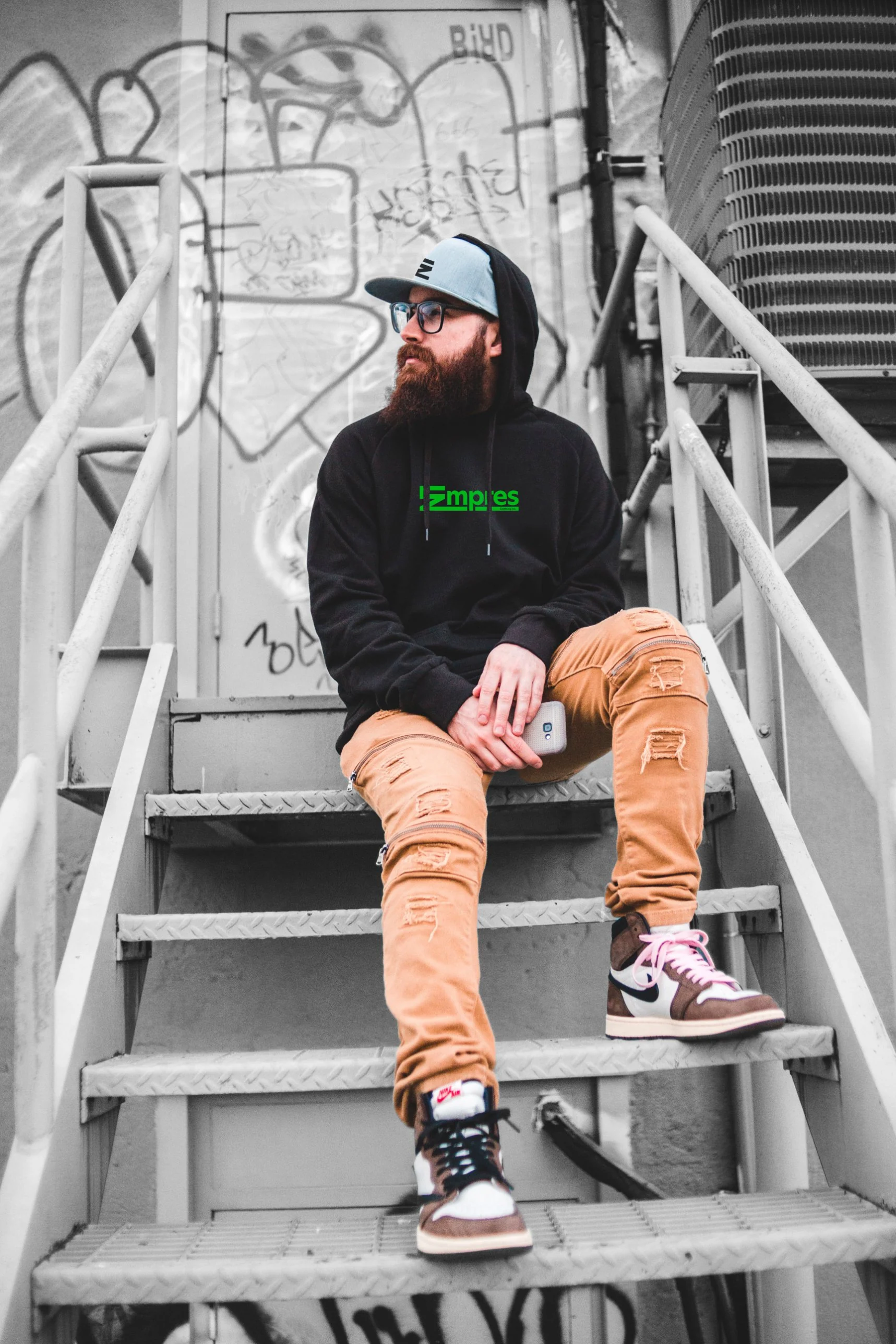 Lookbook photo: bearded man on graffiti-covered metal stairs in a black hoodie with the green Empres wordmark and a grey cap with the E mark